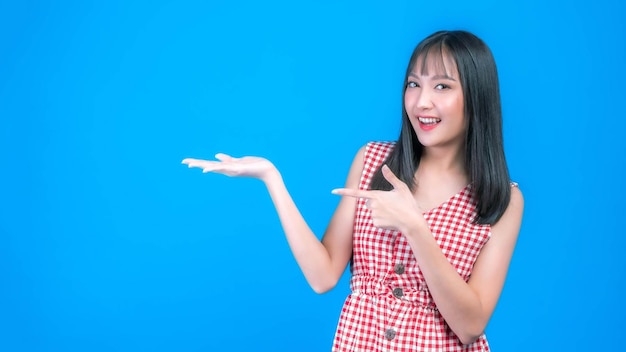 Beautiful beauty Asian woman cute girl with bangs hair style in red dress smile and posing her empty hands with copy space blank space for advertising isolated on blue background