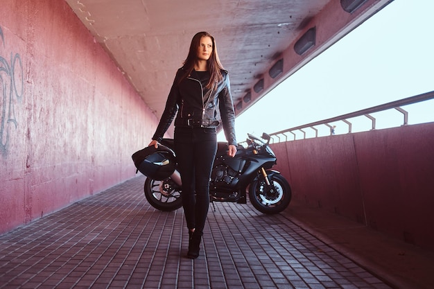 A beautiful biker girl wearing black leather jacket holding helmet next superbike walks on a sidewalk inside the bridge.