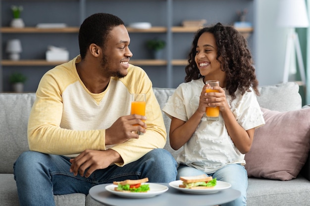 Beautiful black family father and daughter having snack at home, eating healthy fresh sanwiches and drinking orange juice, having pleasure conversation, sitting on couch in living room