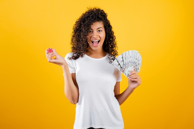 Beautiful black girl with poker chip from online casino and dollars money isolated over yellow