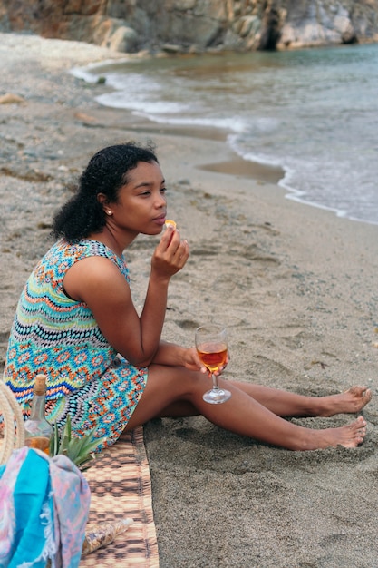 A beautiful Black woman drinks wine on the beach, looking at the horizon.