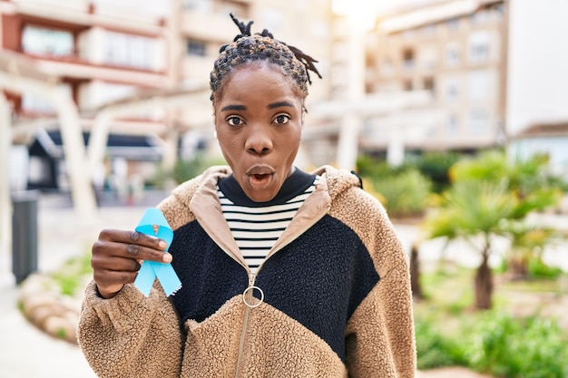 Beautiful black woman holding blue ribbon scared and amazed with open mouth for surprise disbelief face