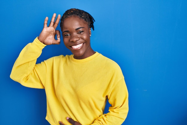 Beautiful black woman standing over blue background smiling positive doing ok sign with hand and fingers successful expression