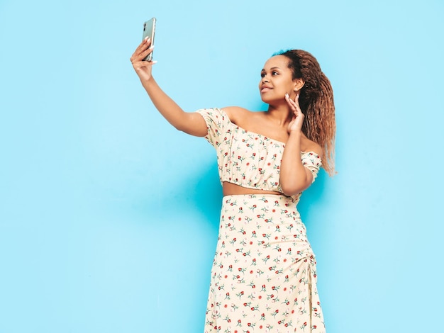 Beautiful black woman with afro curls hairstyle Smiling model dressed in summer dress Sexy carefree female posing near blue wall in studio Tanned and cheerful Taking selfie