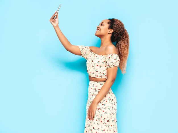 Beautiful black woman with afro curls hairstyle Smiling model dressed in summer dress Sexy carefree female posing near blue wall in studio Tanned and cheerful Taking selfie