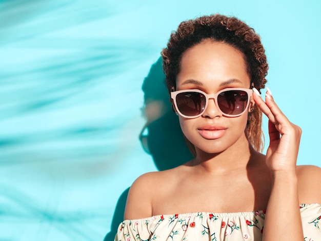 Beautiful black woman with afro curls hairstyle Smiling model dressed in summer hipster clothes Sexy carefree female posing near blue wall in studio Tanned and cheerful In sunglasses