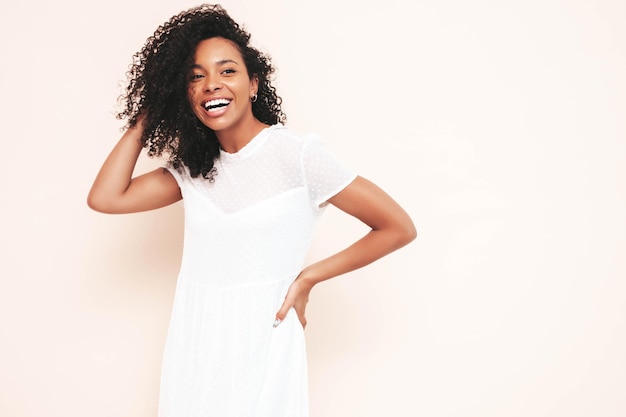 Beautiful black woman with afro curls hairstyle Smiling model dressed in white summer dress Sexy carefree female posing near wall in studio Tanned and cheerful Isolated