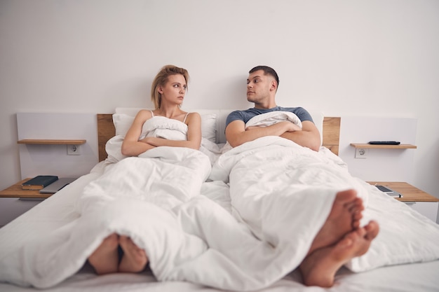 Beautiful blond female and brunette male  looking angrily while lying in bed