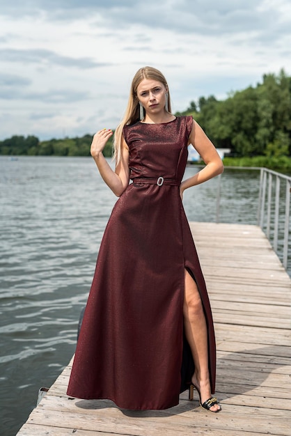 Beautiful blond girl in a long dark red dress posing on the pond romantic