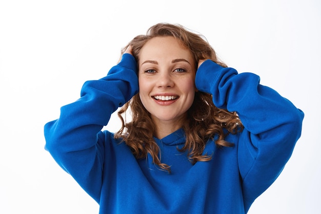 Beautiful blond girl touching her fair curly hair smiling pleased concept of haircare and cosmetics using shampoo or conditioner standing over white background