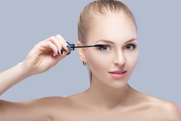 beautiful blond woman applying makeup on face on gray background. perfect makeup for blue eyes. the brush of mascara