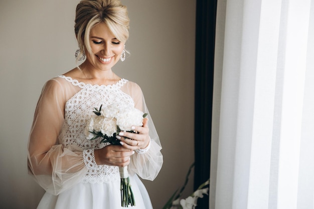 Beautiful blonde bride smiling wearing white wedding dress and holding bouquet indoors