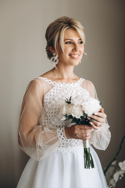 Beautiful blonde bride smiling wearing white wedding dress and holding bouquet indoors