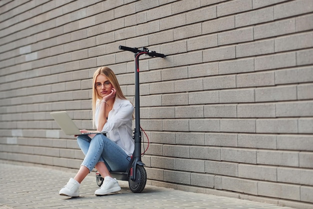 Beautiful blonde in casual clothes sitting on electric schooter outdoors at sunny daytime