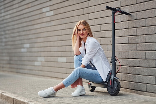 Beautiful blonde in casual clothes sitting on electric schooter outdoors at sunny daytime