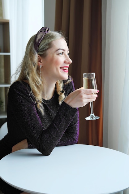 A beautiful blonde girl in a dress holds a glass of champagne in her hands smiles and looks out the window