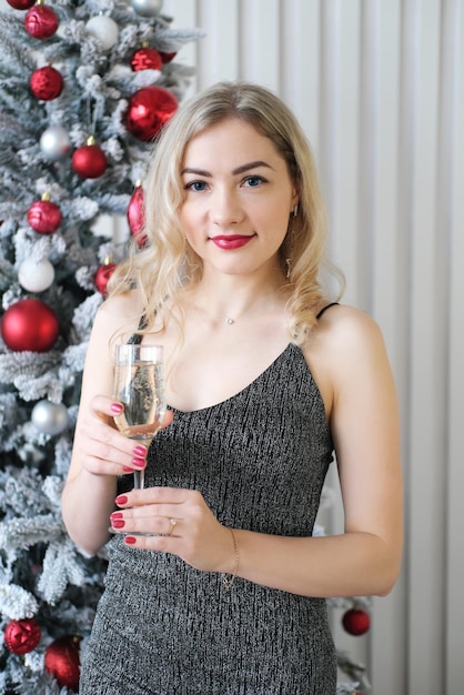 A beautiful blonde girl in a gray shiny dress stands against the backdrop of a Christmas tree with a glass of champagne in her hands