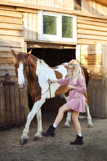 A beautiful blonde girl is playing with his a red and white horse in the stable in full growth