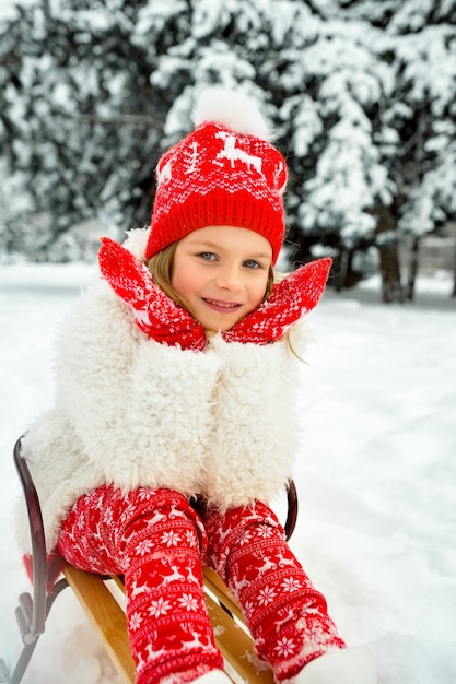 Beautiful blonde girl in a red hat and knitted mittens sits in a sled against snowy park