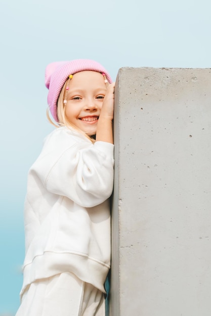 beautiful blonde girl in a white tracksuit and a pink hat climbs on a concrete fence against the backdrop of a city landscape