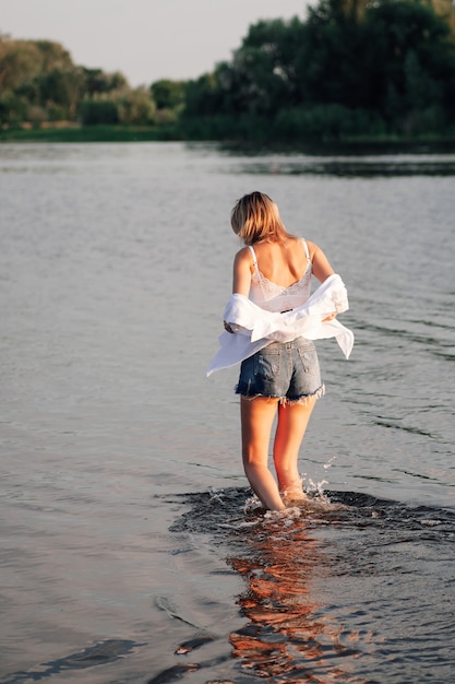 A beautiful blonde is standing with her back to the sunset a young woman in white clothes and denim ...