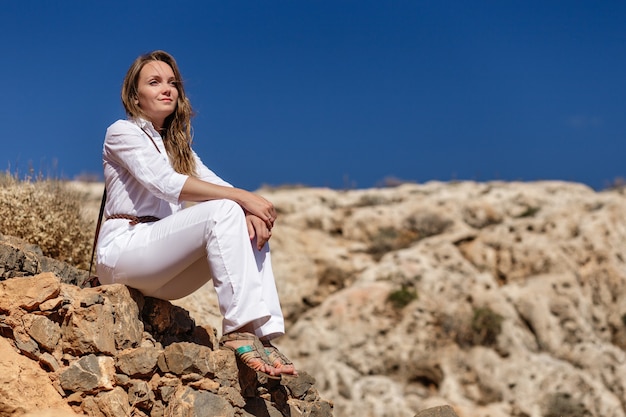 A beautiful blonde in white pants and a blouse is sitting among the stones of gramvousa island