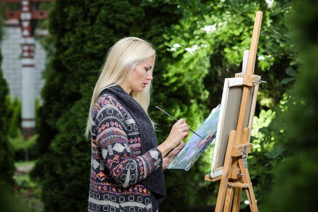 Beautiful blonde woman artist with a brush in her hand draws on canvas in the nature.