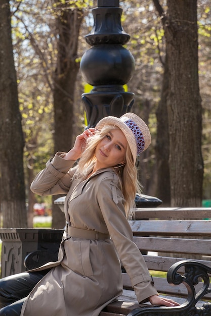 Beautiful blonde woman in a raincoat and hat sits on a bench