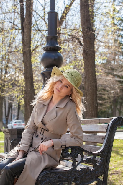 Beautiful blonde woman in a raincoat and hat sits on a bench