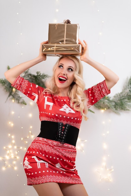 Beautiful blonde woman in a red Christmas dress poses against the background of Christmas tree branches and a glowing garland Holding a gift in his hands Celebration of new year christmas