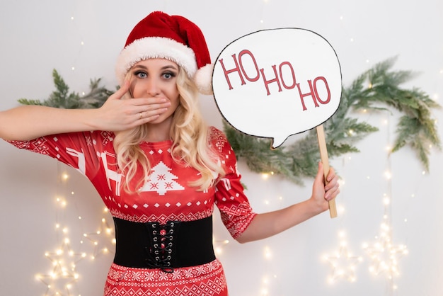 Beautiful blonde woman in a red Christmas dress poses against the background of Christmas tree branches and a glowing garland Holds a sign in his hand that says HoHoHo