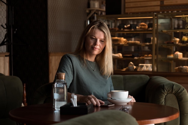 Beautiful blonde woman sits in coffeehouse and bakery and drinks tea or coffee and using smart phone