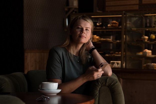 Beautiful blonde woman sits in coffeehouse and bakery and drinks tea or coffee and using smart phone