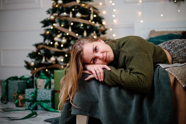 A beautiful blonde woman sits near a Christmas tree in a New Year's atmosphere and rejoices The woman is in a bright comfortable bedroom beautifully decorated for Christmas