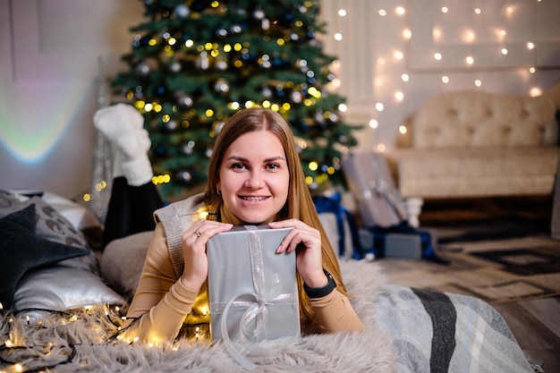 A beautiful blonde woman sits near a Christmas tree in a New Year's atmosphere and rejoices The woman is in a bright comfortable bedroom beautifully decorated for Christmas