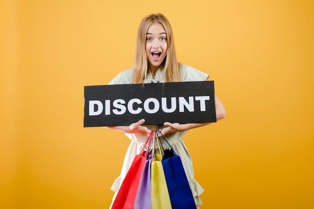 Photo beautiful blonde woman smiling and screaming with discount sign and colorful shopping bags isolated over yellow