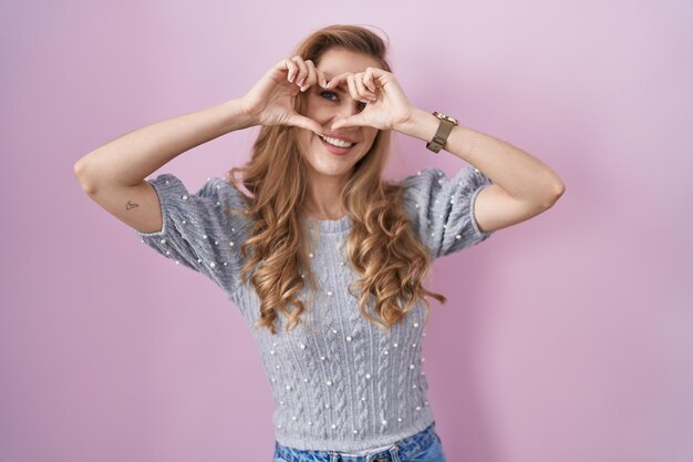 Beautiful blonde woman standing over pink background doing heart shape with hand and fingers smiling looking through sign