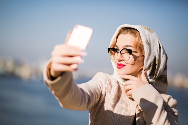 Beautiful blonde woman taking selfie on smart phone. Woman wears shawl and glasses