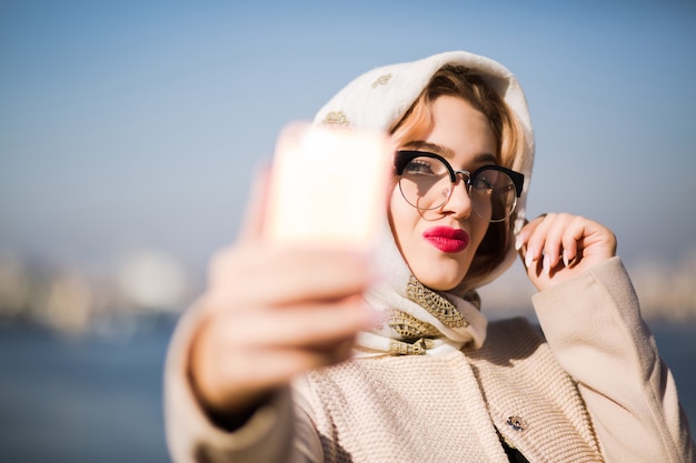 Beautiful blonde woman tourist taking selfie on smartphone. Woman wears shawl and glasses