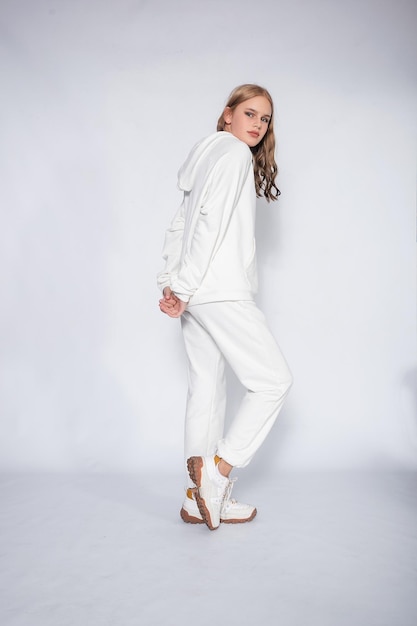 Beautiful blonde woman wear of white set of tracksuit isolated on gray background blonde girl in a white tracksuit stands on a white background