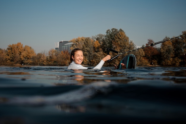 Beautiful blonde woman in the white swimsuit holding a green wakeboard in the river water