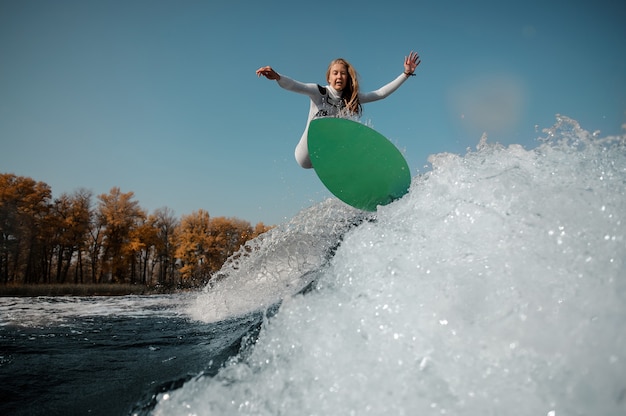 Beautiful blonde woman in the white swimsuit jumping on the green wakeboard on the bending knees