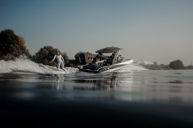 Beautiful blonde woman in the white swimsuit riding on the wakeboard holding a rope of the motorboat