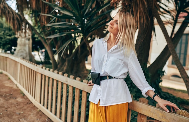 Photo beautiful blonde woman with closed eyes enjoying the warm weather in the park modern stylish female wearing white shirt and yellow skirt relaxing in the summer time in the city park natural lighting