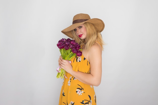 Beautiful blonde woman in yellow floral dress and straw hat holding bouquet of purple tulips