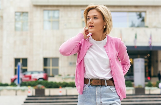 Beautiful blonde young woman wearing blue jeans white tshirt and pink jacket smiling and taking selfie on smart phone Happy female making self portrait in the city street in spring time