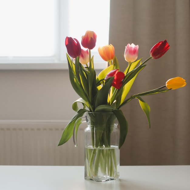 Beautiful bouquet of tulips in glass vase on white table in a cozy room interior Blooming flowers decoration in living room