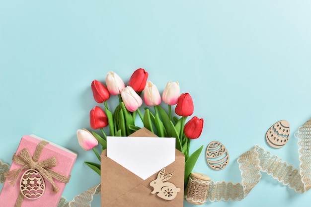 A beautiful bouquet of tulips with a postcard and a gift and wooden Easter eggs on a light blue background Banner for happy Easter Space for copying Flat position top view