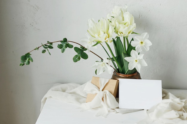 Beautiful bouquet in vintage vase gift box and empty card on rustic wooden table Greeting card mockup and stylish white spring tulips Happy Mothers day Women's day Space for text