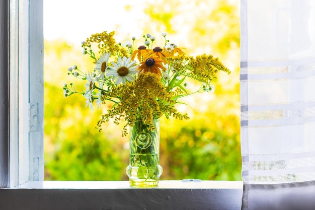 A beautiful bouquet of wildflowers on a white windowsill by an open window with a white curtain in a country house on a summer evening